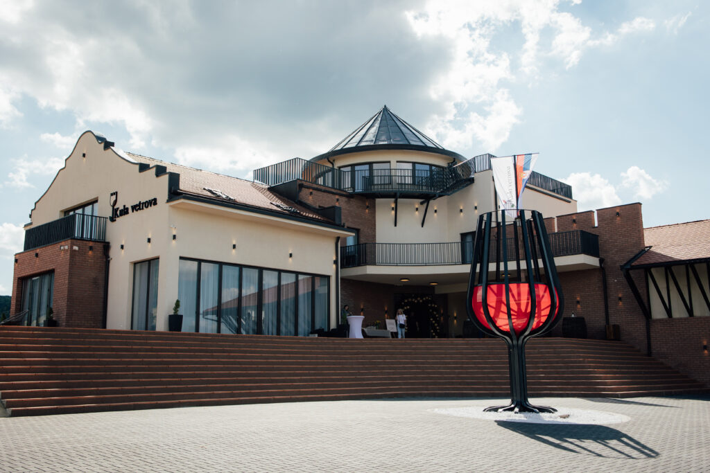 Dnevna fotografija vinarije Kula vetrova sa kružnom kulom, oblikovanim zabatom i širokim ulaznim stepeništem